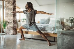 Black man in Warrior II pose in front of his bed at home