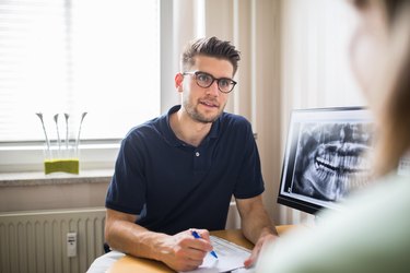 A dentist with short brown hair and glasses talking to an unrecognizable patient about their X-rays