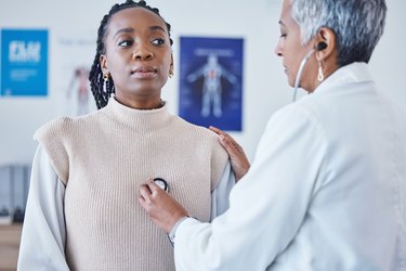 A doctor with short gray and black hair placing a stethoscope on chest of a young patient wearing a beige sweater checking for lung and heart health at hospital