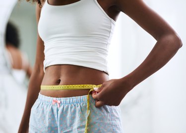 A woman in pajamas measuring her waist with a yellow cloth tape