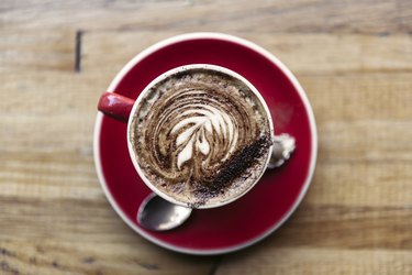 Overhead photo of a mug of coffee with cocoa powder served in a red cup and saucer on a rustic wooden table