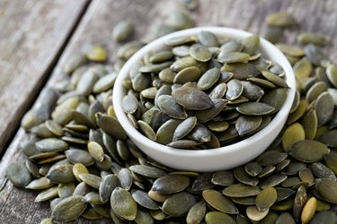 a small white bowl of pumpkin seeds surrounded by more pumpkin seeds on a wooden surface