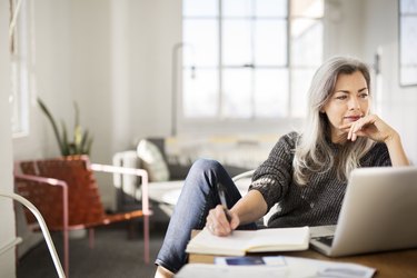 person with long grey hair sitting at a desk and writing in a notepad