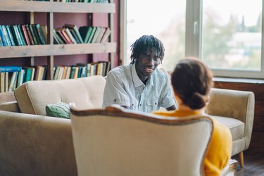 a person with short black dreadlocks wearing a white button-down shirt and sitting on a beige couch in a therapists office